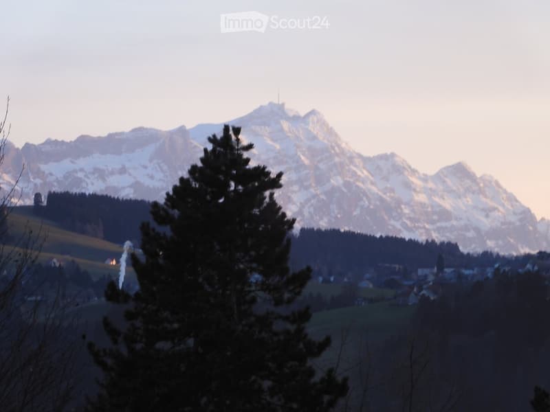 A scenic view of snow-capped mountains in the background, with a small village nestled in the valley below, surrounded by trees and fields.