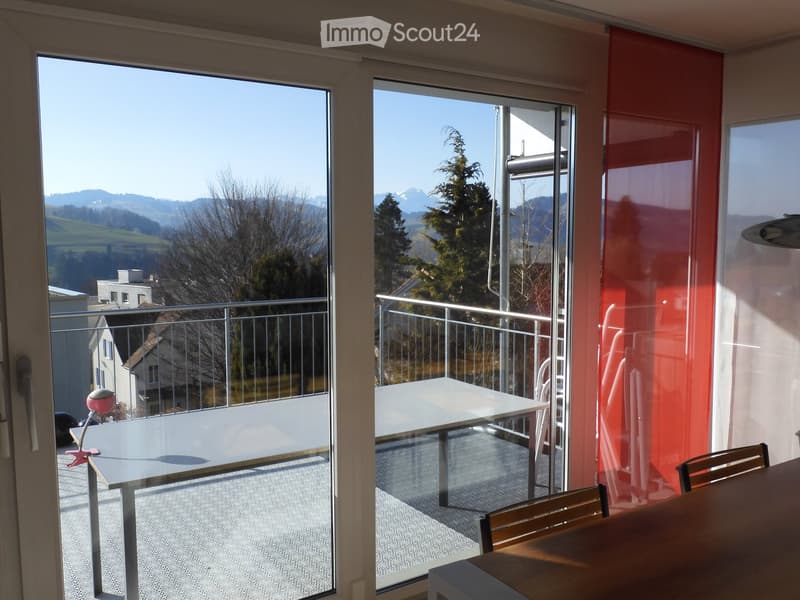 Balcony with a table and chairs, overlooking a scenic mountain view with trees and houses.