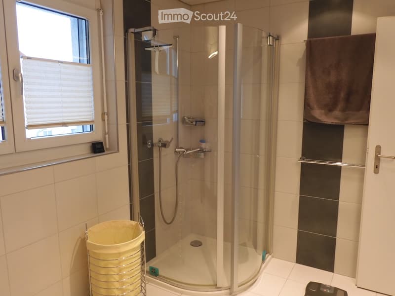 Modern bathroom with a glass-enclosed shower stall, beige tiles, a towel hanging on a rack, and a small trash bin near the shower.