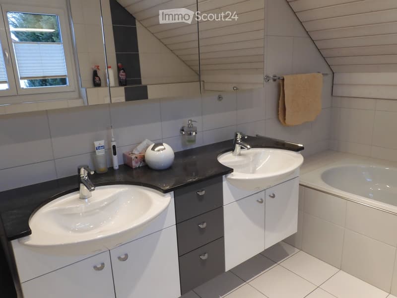 Modern bathroom with double sinks, a bathtub, a large mirror, and a towel hanging on the wall. The countertop has various toiletries and a soap dispenser.