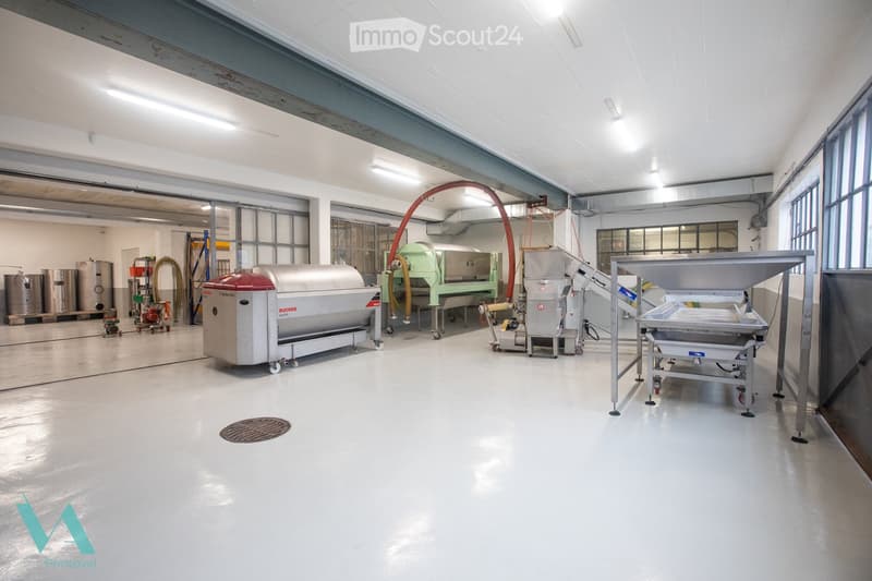 Industrial space with large machinery, cylindrical tanks, conveyor belt, and various pipes. The floor is white and the ceiling has exposed beams and lighting.