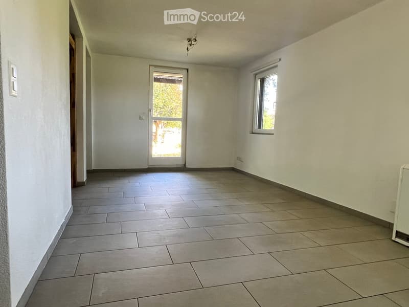 Empty room with tiled floor, white walls, and a large window with a view of greenery. The room has a door leading outside and a small window on the left side.
