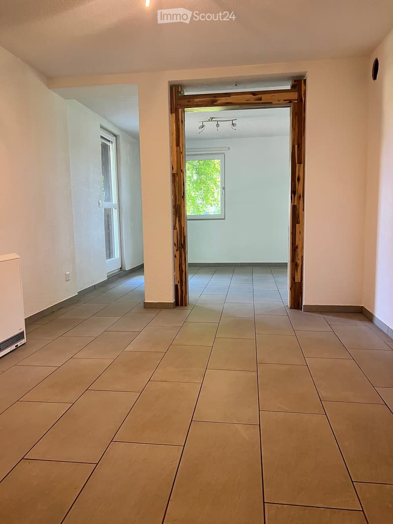 Empty hallway with tiled floor, light-colored walls, and wooden door frame leading to another room with a window.