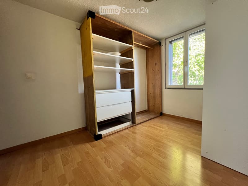 Empty room with wooden flooring, white walls, and a built-in wooden shelving unit with drawers. A window with white frames allows natural light to enter.
