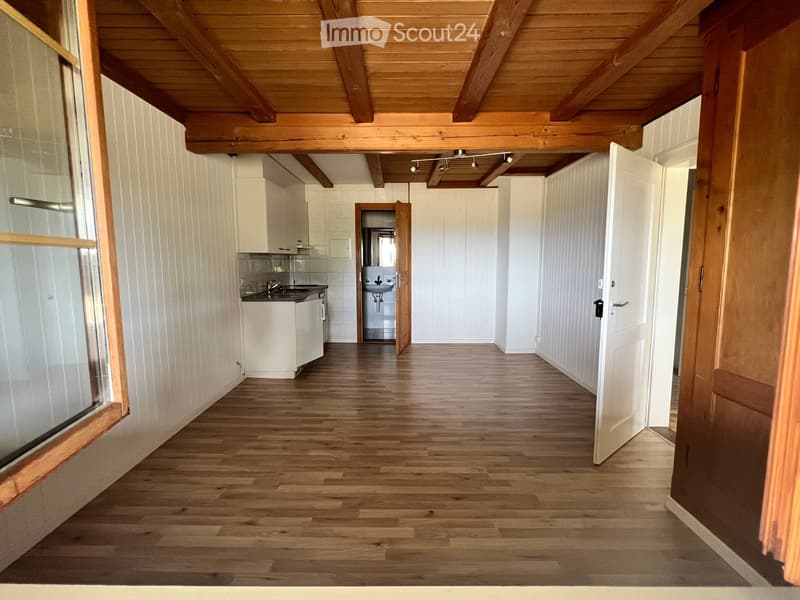 Empty wooden-floored hallway with white walls, exposed wooden beams on the ceiling, and doors leading to a kitchen and bathroom.