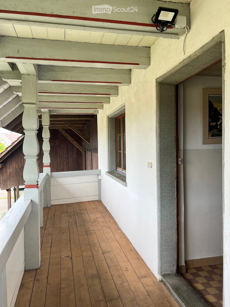 Wooden hallway with white painted columns, red trim, and a small window. A painting is visible on the wall at the end of the hallway.