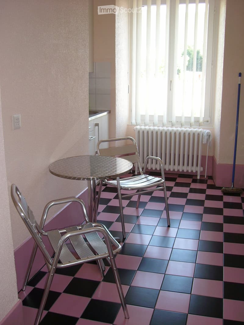 A small room with a table and chairs, pink and black checkered floor, white radiator, and a window with white blinds.