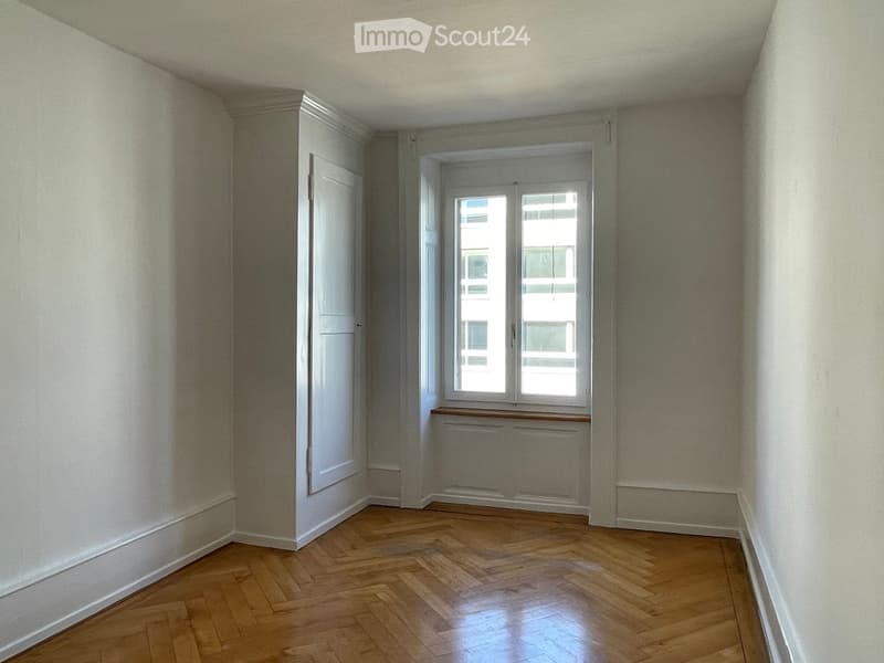 Empty room with white walls, wooden parquet floor, and a large window providing a city view.