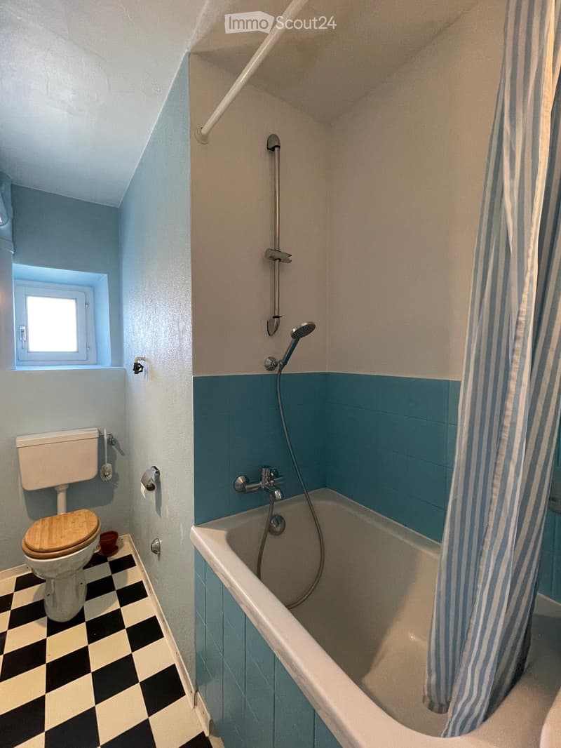 Bathroom with blue and white walls, checkered floor, bathtub, shower head, toilet, small window.