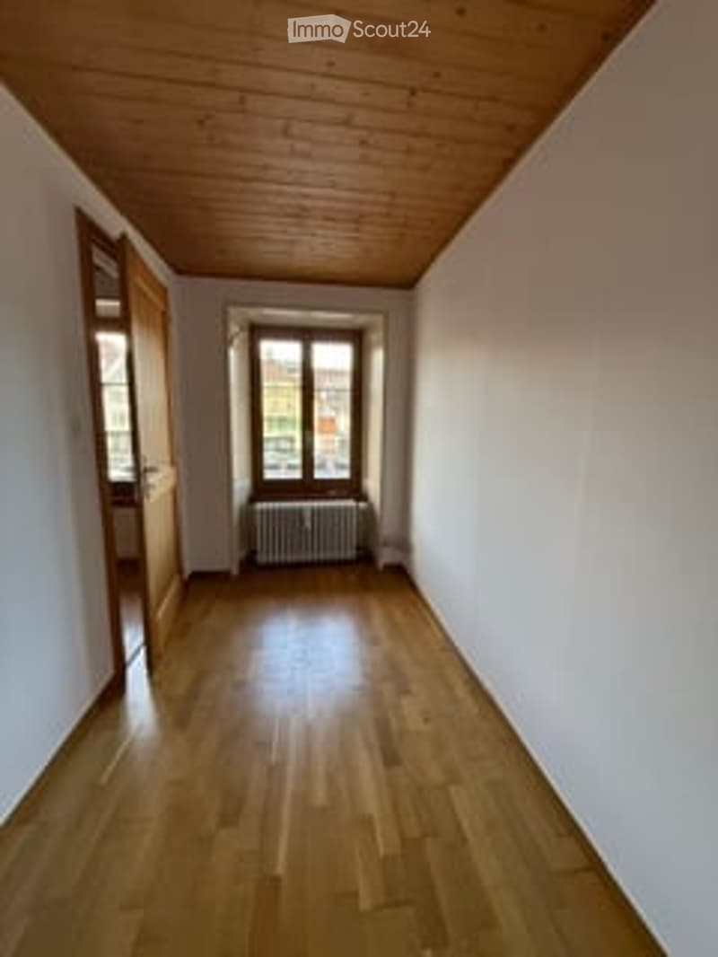 Unfurnished Room. Empty room with wooden flooring, white walls, and a single window with a radiator beneath it. The ceiling is wooden, and there is a door on the left side.