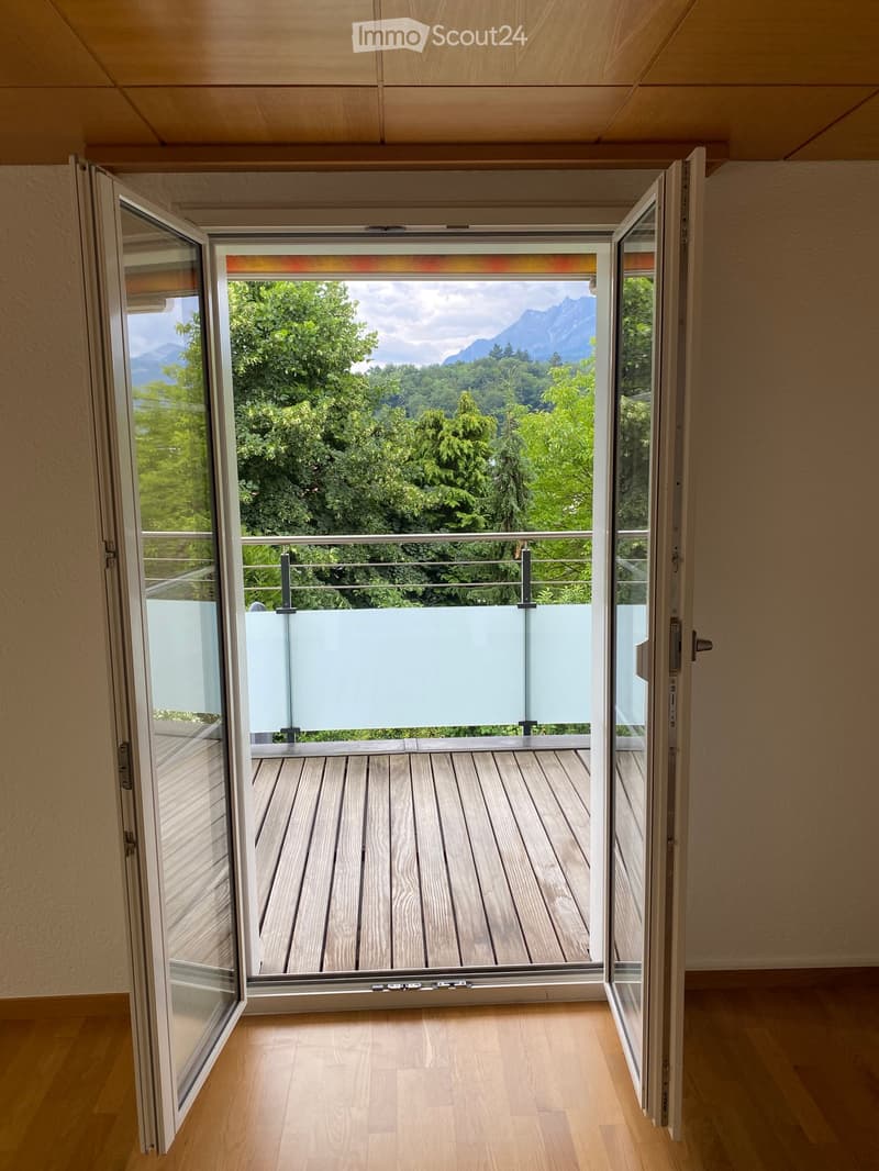 Open sliding doors lead to a wooden deck with a glass railing, offering a scenic mountain and forest view.