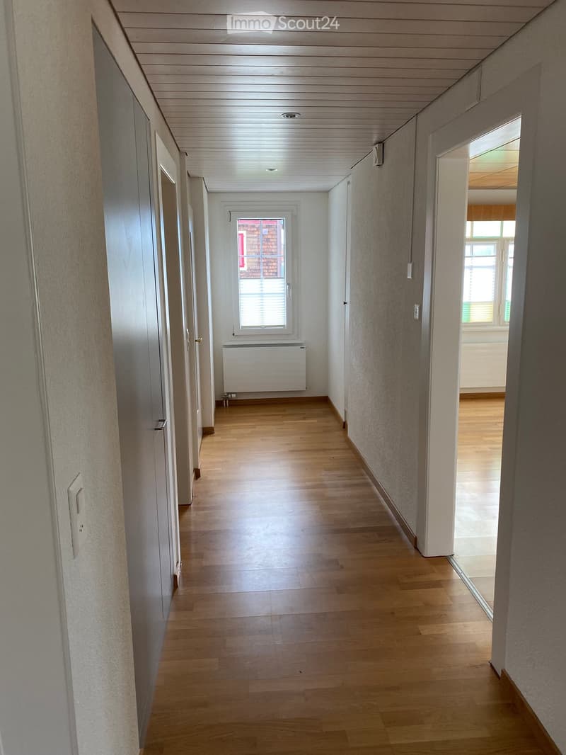 Empty hallway with wooden flooring, white walls, and several doors. A window with blinds is visible at the end of the corridor, along with a radiator beneath it.