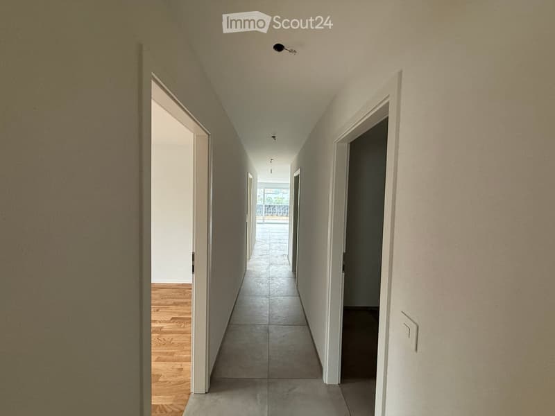 Corridor / Hallway. A long, narrow hallway with light-colored walls, wooden flooring on one side, and tiled flooring on the other. Several doors are visible along the walls, and the ceiling has recessed lighting.