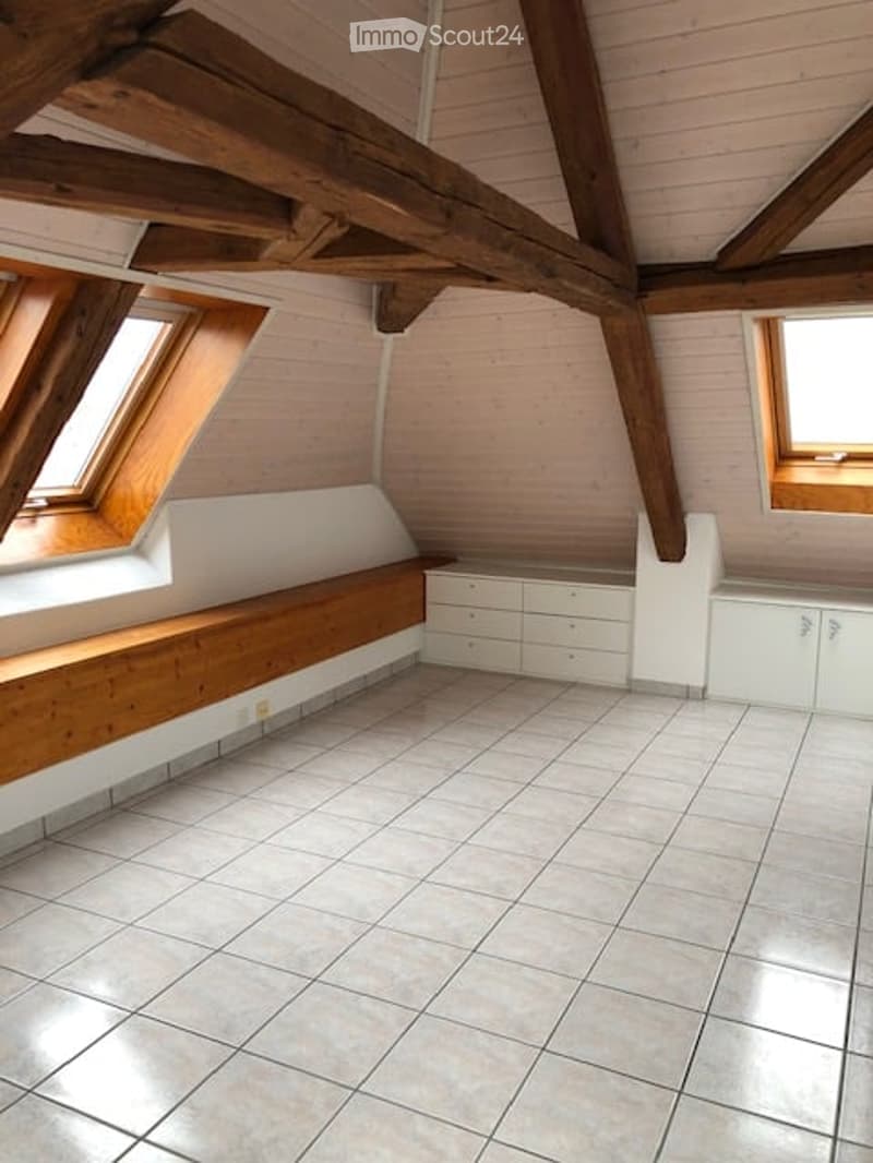 An attic room with exposed wooden beams, white paneled walls, tiled floor, built-in bench seating, and built-in cabinets. The room has skylight windows allowing natural light to enter.
