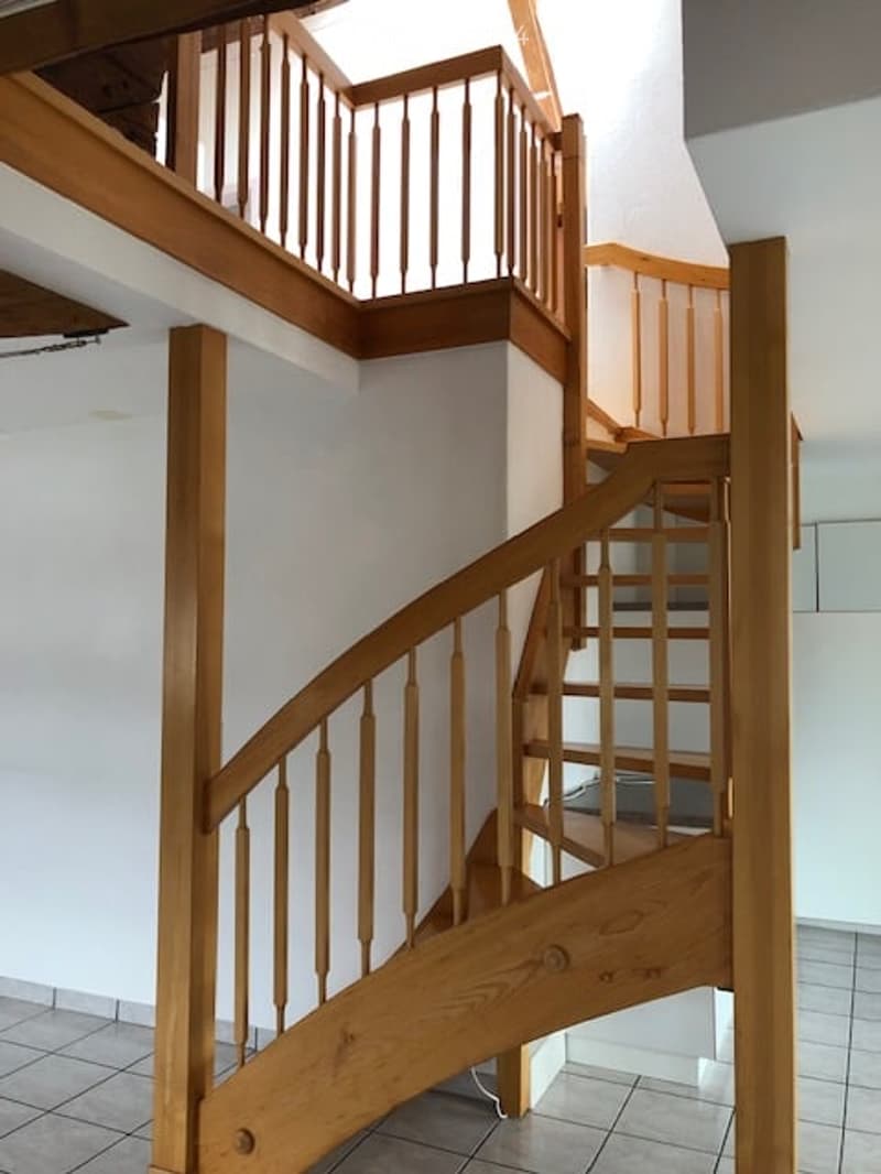 Spiral staircase with wooden handrails and steps, white walls, tiled floor, leading to an upper level.