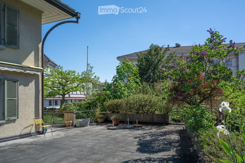 A sunny garden patio with a stone floor, potted plants, a metal railing, and a yellow chair. The background shows a house with shutters and neighboring houses with trees and flowering shrubs.