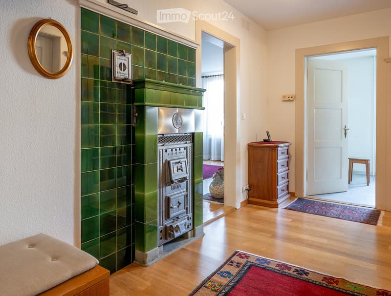 Hallway with wooden flooring, green tiled fireplace, small bench, and open doors leading to other rooms.