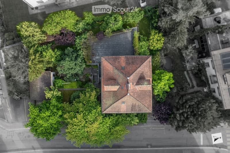 Aerial view of a residential house with a red-tiled roof, surrounded by lush green trees and a well-maintained garden. The house has a driveway and is situated in a quiet neighborhood.