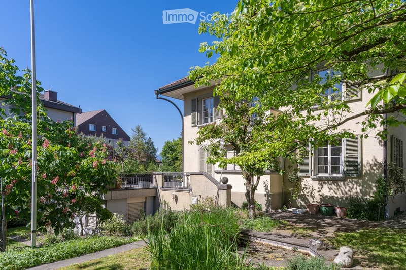 A two-story residential building with a well-maintained garden, featuring a balcony, large windows, and lush greenery.