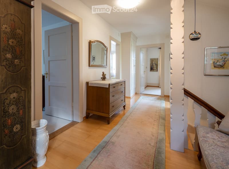 Elegant hallway with wooden flooring, a patterned rug, a small wooden cabinet with drawers, a mirror, and framed artwork on the walls.