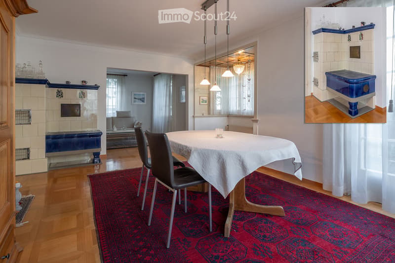Dining area with a wooden table, chairs, and a red rug. Adjacent living area features a sofa, fireplace, and large windows with curtains.