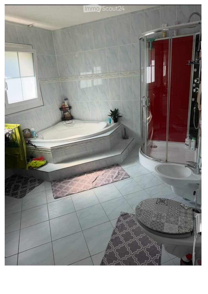 Modern bathroom with a large bathtub, separate shower, sink, toilet, and decorative rugs. Features light blue tiles, a window, and a small plant on the edge of the tub.