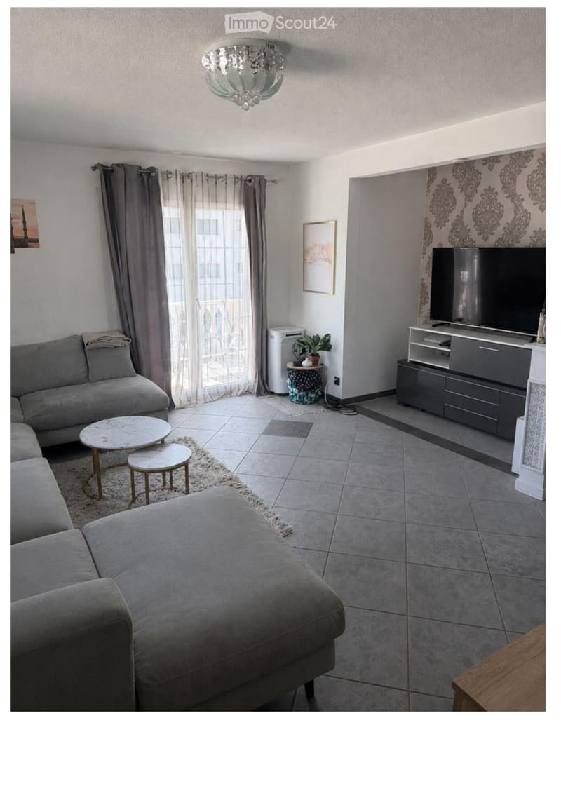 Modern living room with grey sectional sofa, ottoman, coffee table, two small stools, TV stand with flat-screen TV, floor lamp, picture frame, small plant, and large window with curtains.