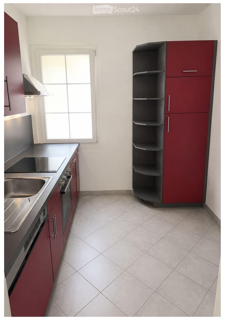 Modern kitchen with red cabinets, stainless steel sink, oven, and shelves. The space is well-lit with a large window and tiled flooring.