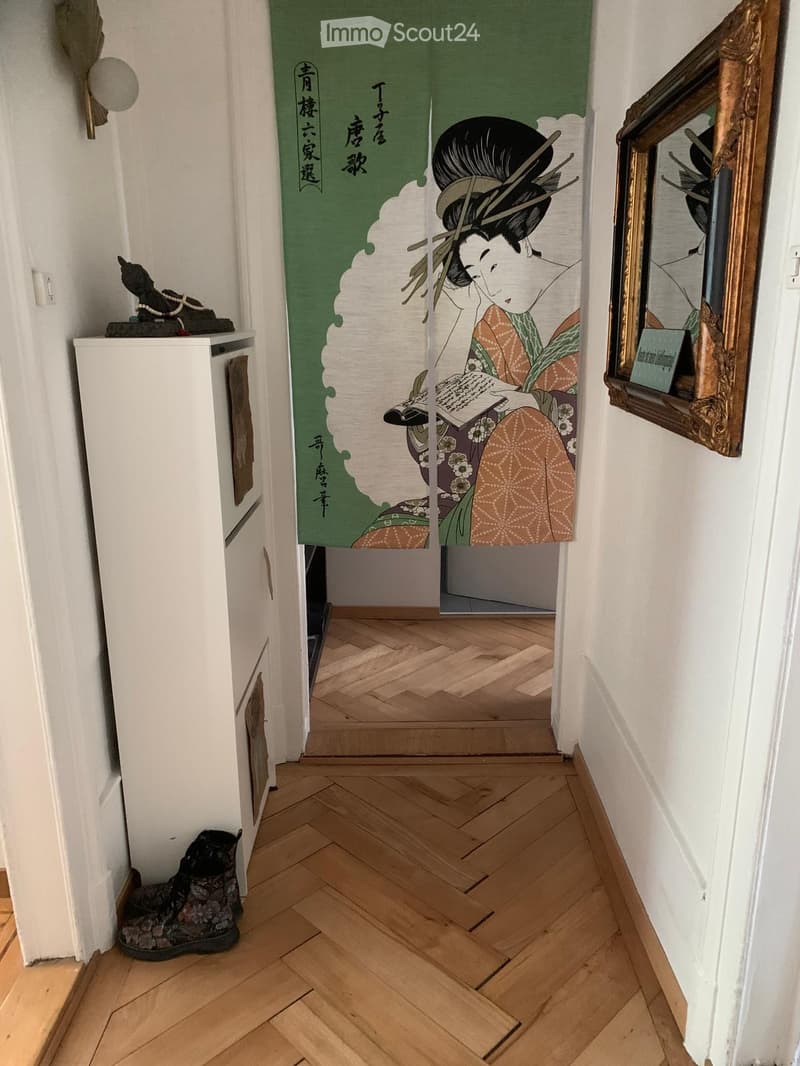 A narrow hallway with wooden herringbone flooring, white walls, a large framed artwork, and a small cabinet with a decorative item on top.
