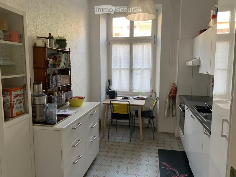 The kitchen features white cabinets, a tiled floor, a window with blinds, a small dining table with two chairs, and various appliances and items on the counters.