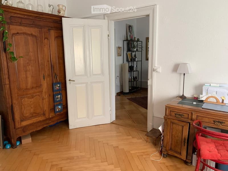 A furnished room with wooden flooring, a large wooden wardrobe, a desk with a lamp, a red chair, and an open door leading to another room with a shelf and a radiator.