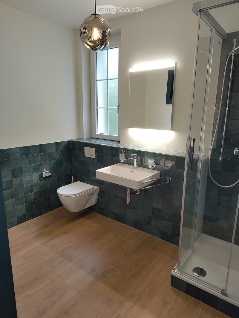 Modern bathroom with dark green tiles, white sink, toilet, and glass-enclosed shower. Features a pendant light and a window with frosted glass.