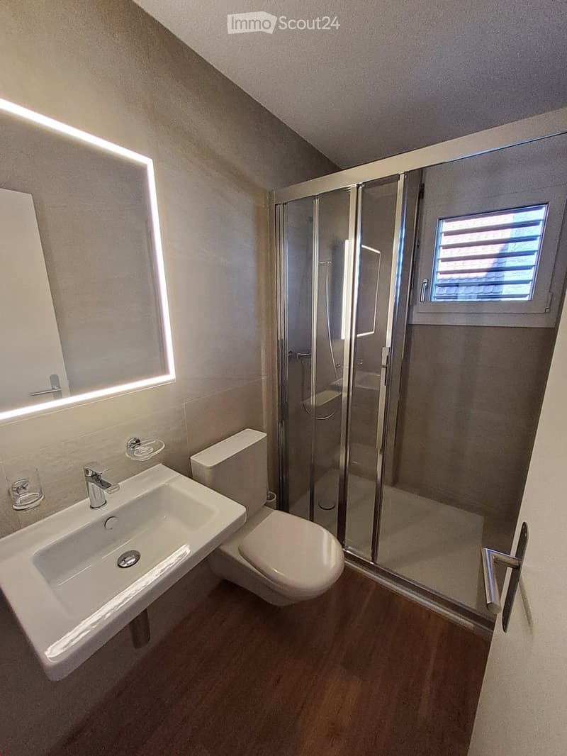 Modern bathroom with a white sink, toilet, glass-enclosed shower, and a small window above the shower. The walls are tiled, and the floor is wooden.