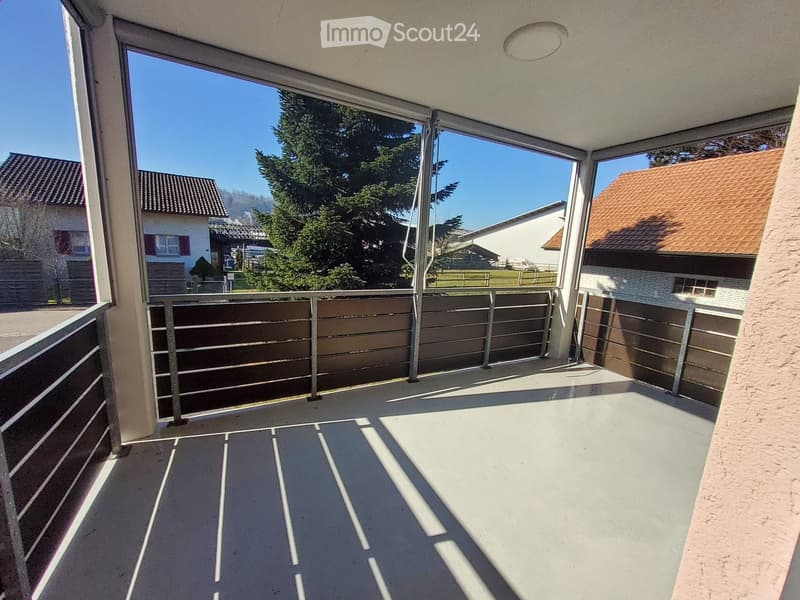 Balcony with metal railing, concrete floor, and large windows overlooking a residential area with trees and houses.