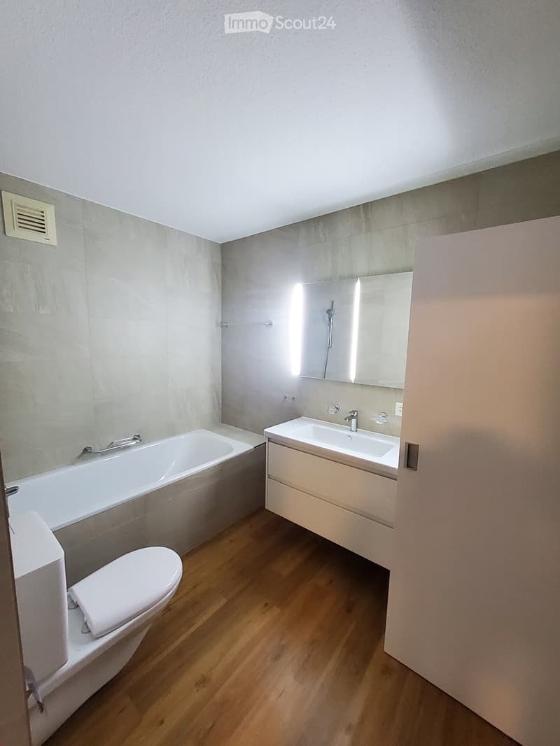 Modern bathroom with a bathtub, toilet, sink, and mirror. Light grey tiles and wooden flooring.