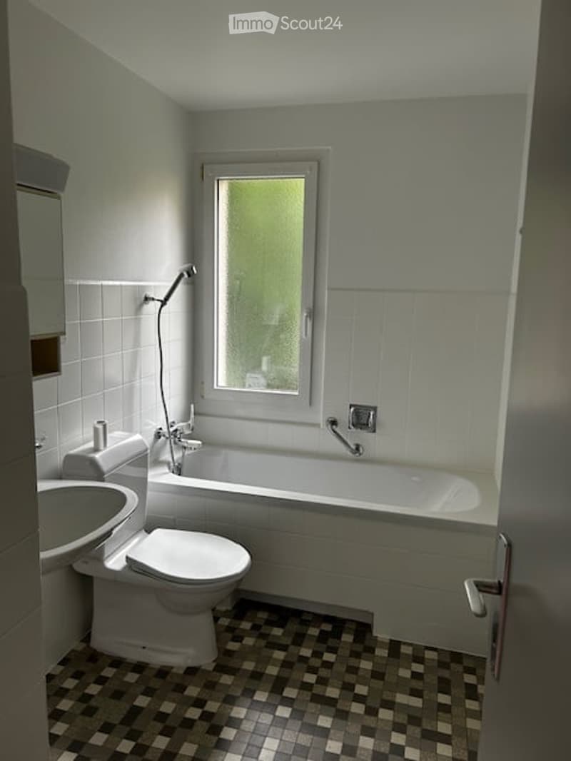 Bathroom with a toilet, sink, bathtub, and a window above the tub. The walls are tiled, and the floor has a checkered tile pattern.