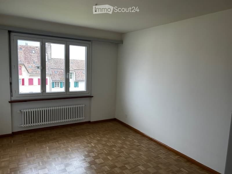 Completely empty room with wooden flooring, white walls, and a large window offering a view of neighboring buildings.