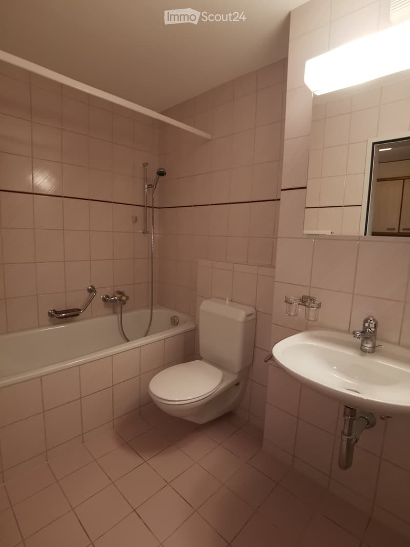 Bathroom with beige tiles, bathtub, toilet, sink, and mirror. Includes a handheld showerhead and a cup on the wall.
