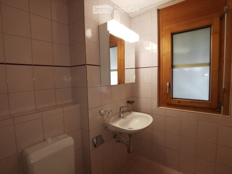 Bathroom with beige tiled walls and floor, a white sink with a chrome faucet, a mirror with built-in lighting, and a toilet. A frosted window with wooden frame is on the right side.