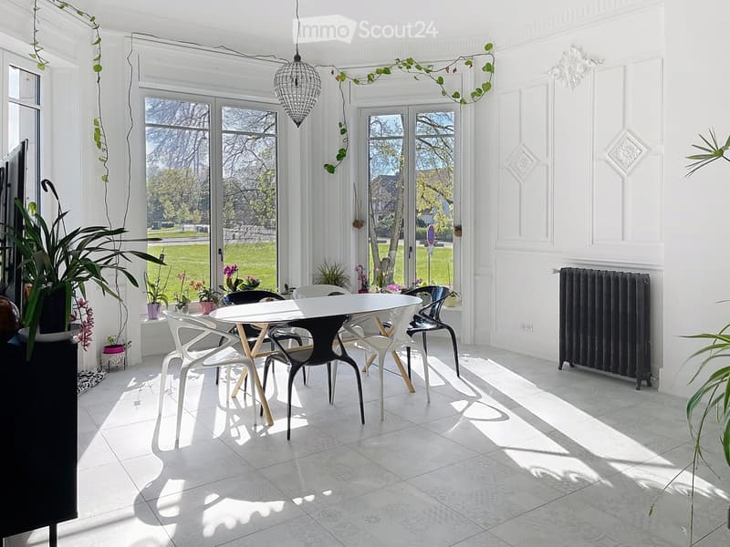 Bright, airy room with large windows, white walls, and a modern dining setup featuring a white table and colorful chairs. Potted plants and a radiator are visible.