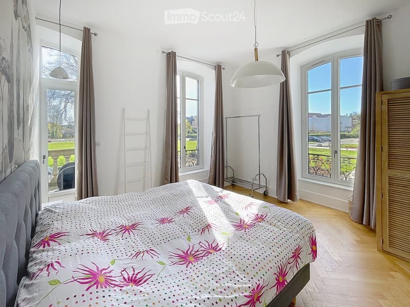 Bright bedroom with wooden flooring, large windows with curtains, a bed with a floral duvet, and a hanging lamp.