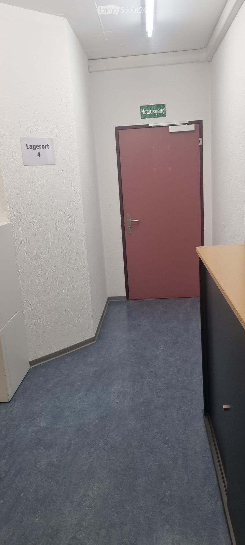 A narrow hallway with blue carpeted flooring, white walls, and a closed maroon door labeled 'Notausgang' (emergency exit). A sign on the left wall reads 'Lagerort 4' (storage location 4).