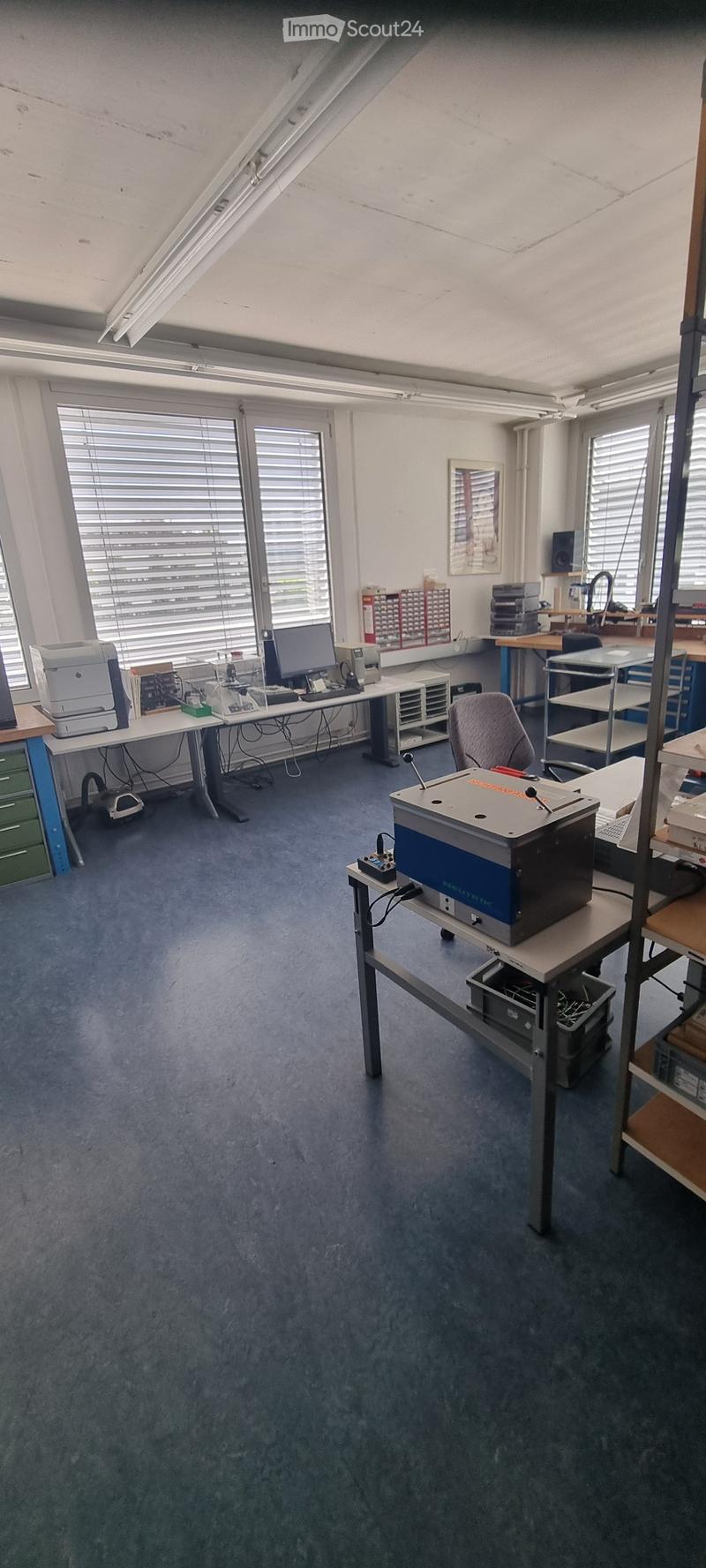 Office space with multiple workstations, desks, chairs, printers, and shelves. Large windows with blinds provide natural light.