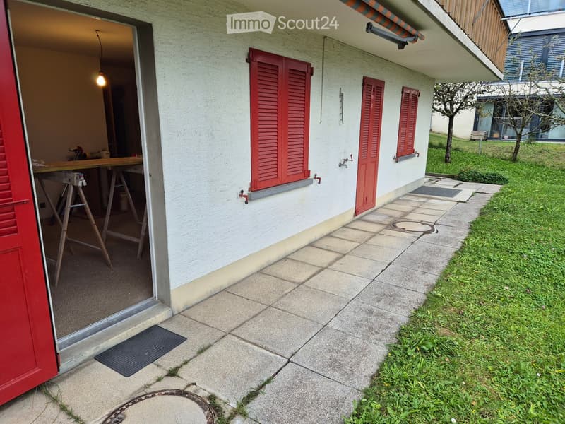 Side view of a house with red shutters, a paved pathway, and a partially visible garage with a workbench inside.