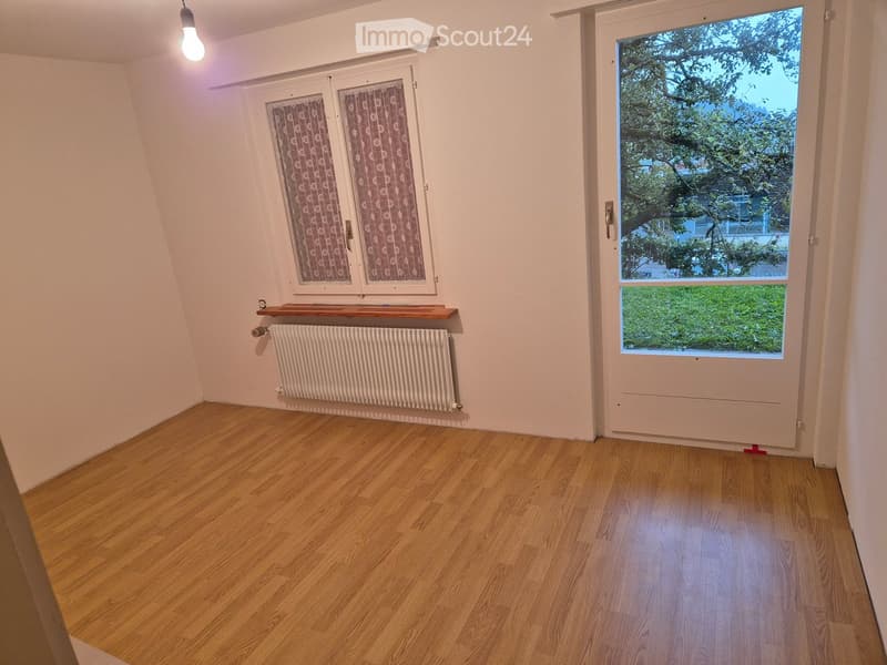 Empty room with wooden flooring, white walls, a radiator, and a window with patterned curtains. A door leads to an outdoor area with greenery visible.