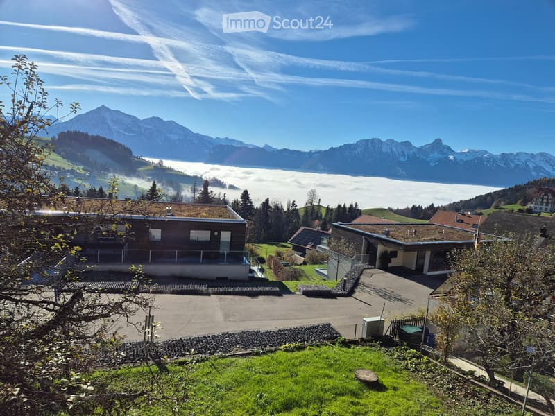 Modern house with a flat roof and large windows, situated in a scenic valley with mountain views and a clear blue sky.