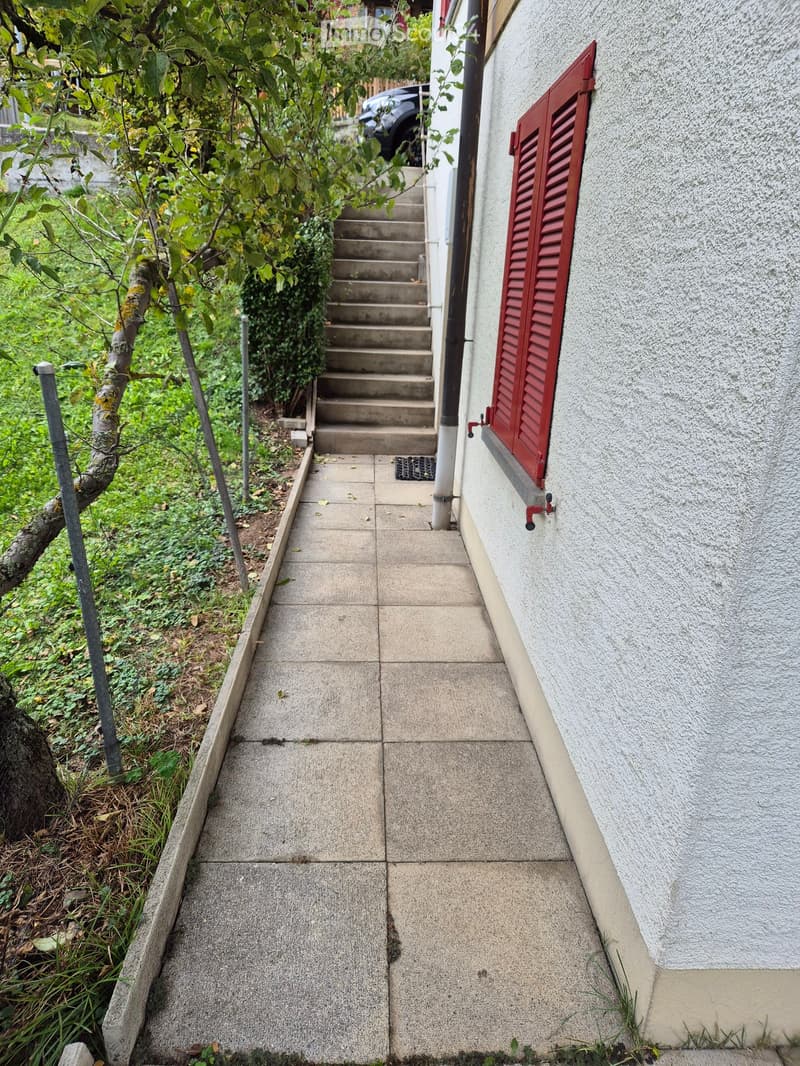 A concrete pathway leading to a raised entrance with steps, featuring a tree and greenery on one side and a building with a red shutter on the other.
