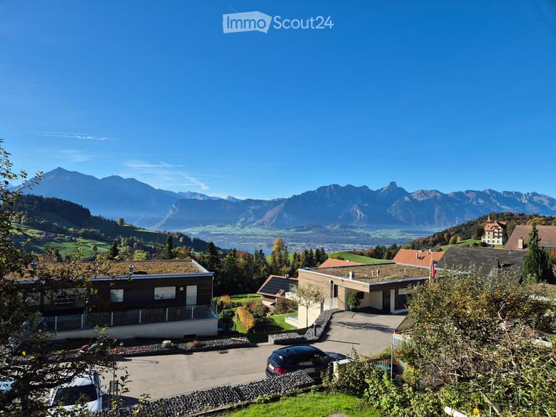 Modern two-story house with a flat roof, large windows, and a parking area, surrounded by greenery with mountain views in the background.