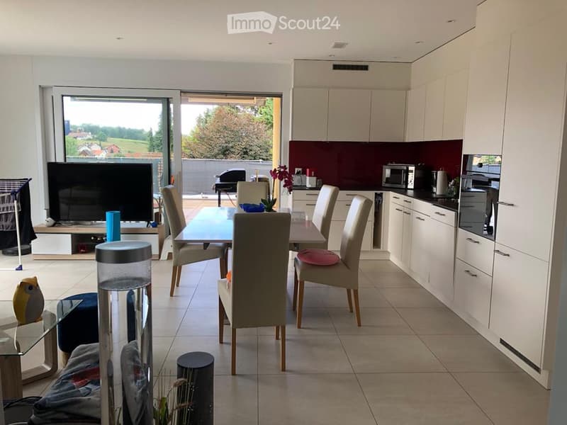 Open-plan kitchen and dining area with modern white cabinets, a dining table with six chairs, a microwave, refrigerator, and a television in the living area. Large windows offer a scenic view of the outdoors.