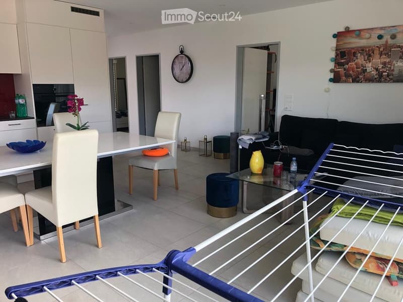 Modern open-plan living and dining area with white dining chairs, a sofa, a glass coffee table, and a clock on the wall.